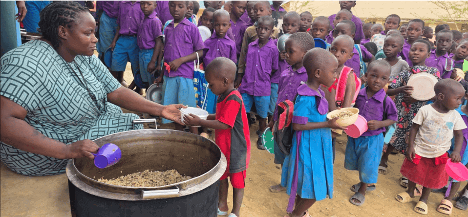 A teacher serves primary school children in Kilifi County, Kenya, as they queue to receive a meal of rice, lentils and dried vegetables through Plan International's school feeding program.